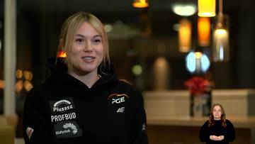 Natalia Maliszewska, polska panczenistka, ubrana w czarną bluzę z logo sponsor&oacute;w, uśmiecha się podczas wywiadu. W tle widoczne są lampy i ekran z inną osobą.