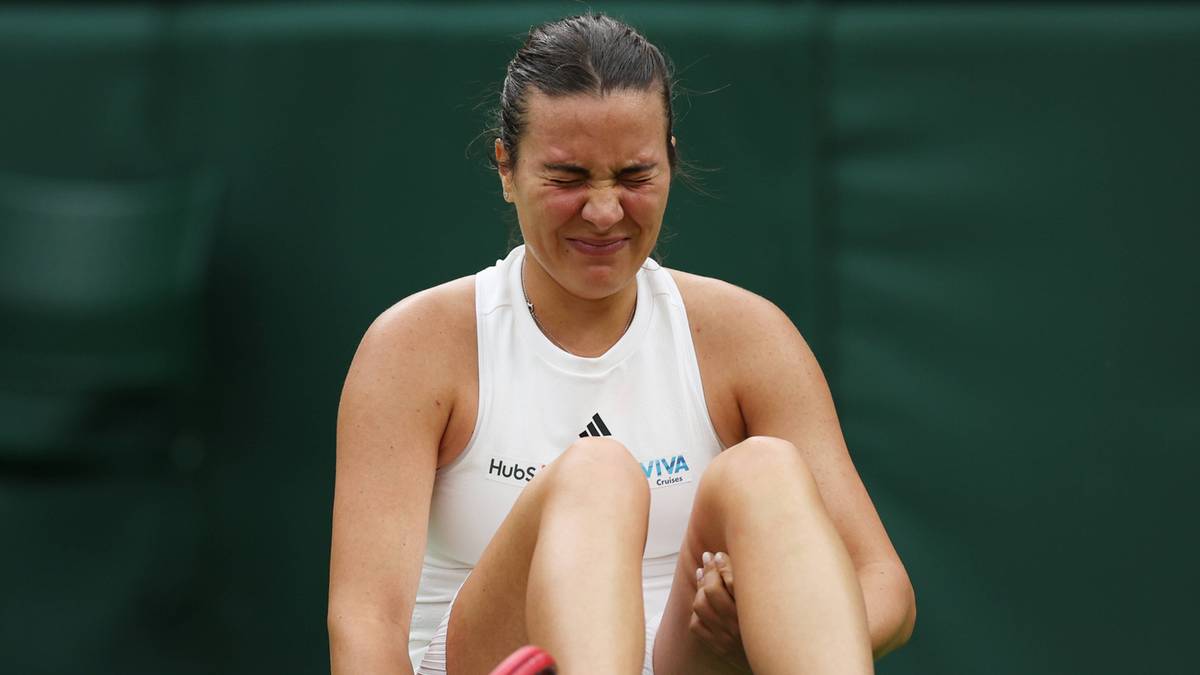 Tenisistka siedzi na trawie z bólem na twarzy, trzymając rakietę i dotykając nogi.