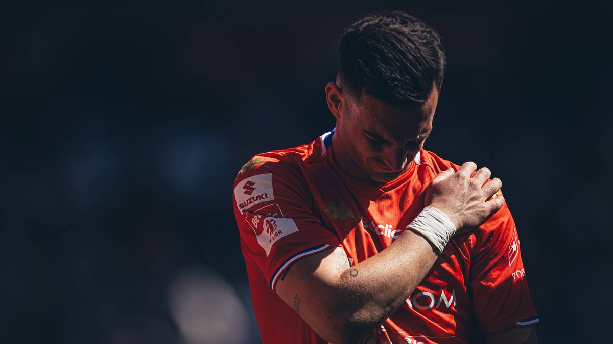 Piłkarz w czerwonej koszulce z ręką na ramieniu, sugerujący b&oacute;l.