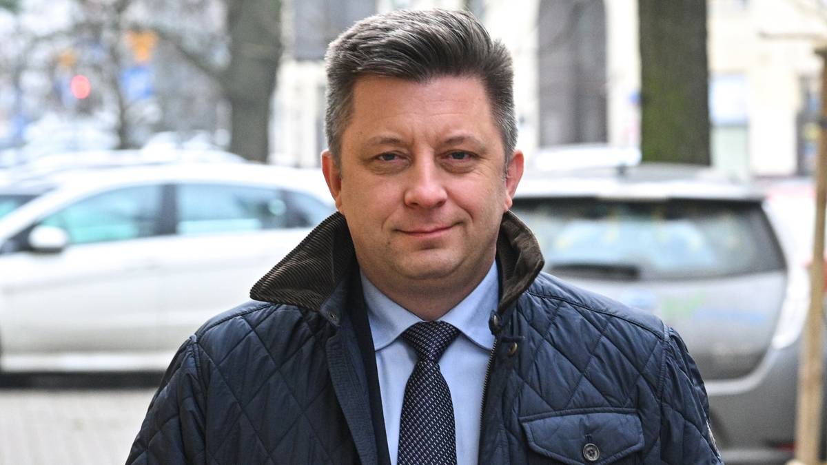 Mężczyzna w średnim wieku, ubrany w ciemną, pikowaną kurtkę, białą koszulę i granatowy krawat, stoi na zewnątrz, z lekkim uśmiechem na twarzy.