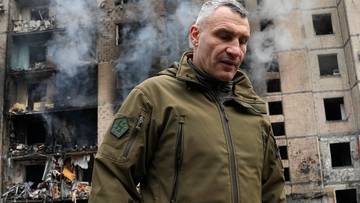 Mer Kijowa Witalij Kliczko stoi przed zniszczonym budynkiem mieszkalnym, z kt&oacute;rego unosi się dym.