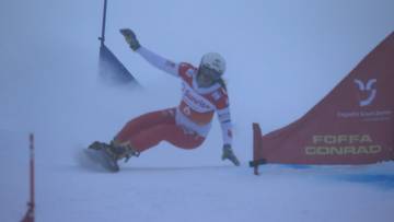 Snowboardzistka w czerwono-białym stroju wykonuje skręt na stoku narciarskim, otoczona śnieżnym pyłem. Widoczna jest pomarańczowa bandę z napisami.