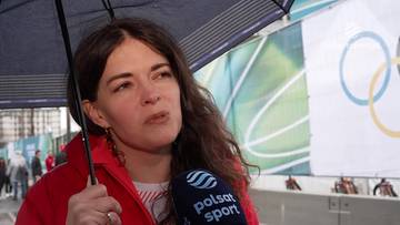 Kobieta z brązowymi włosami trzyma parasolkę, podczas gdy mikrofon Polsat Sport jest skierowany w jej stronę. W tle widoczny baner z symbolami olimpijskimi.