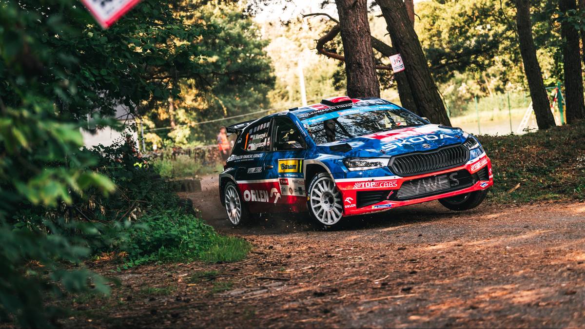 Niebiesko-czerwono-biała rajdowa Skoda Fabia ściga się po piaszczystym, leśnym torze.