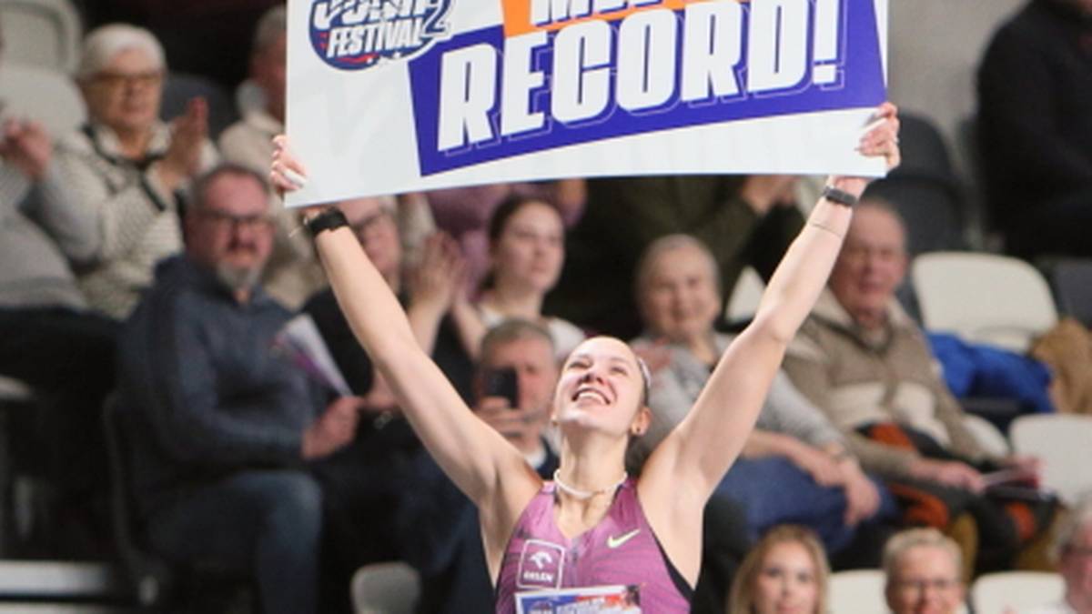 Uśmiechnięta sportsmenka z podniesionymi rękami trzyma baner z napisem "MEETING RECORD!". W tle widownia.
