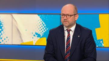 Mężczyzna w garniturze i okularach siedzi, patrząc w bok.