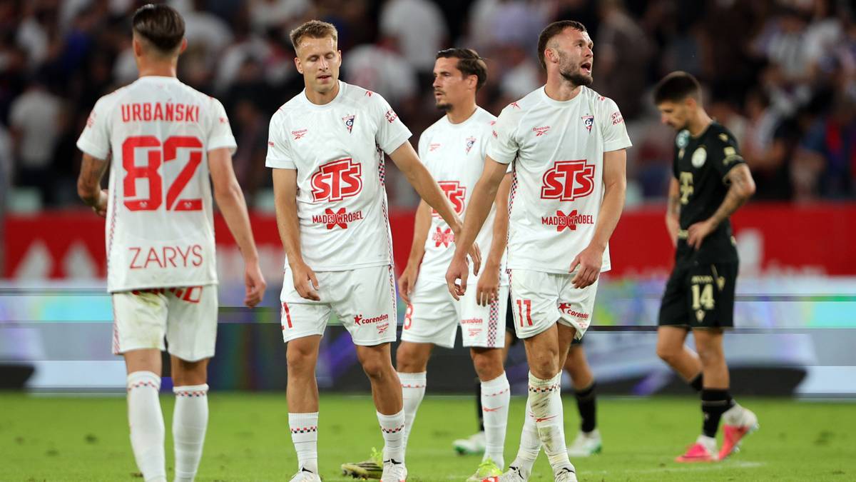 Pięciu piłkarzy w białych strojach z czerwonymi numerami i napisami, stojących na boisku piłkarskim.