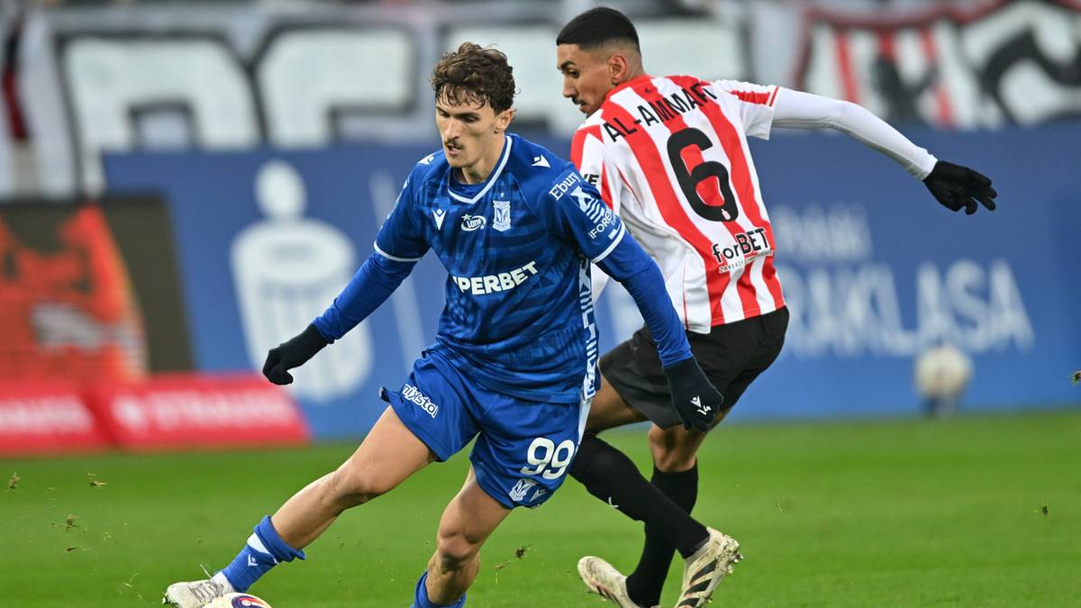 Dwóch piłkarzy grających w piłkę nożną na boisku. Jeden zawodnik w niebieskim stroju z numerem 99 biegnie z piłką, a drugi w białej koszulce w czerwone pasy z numerem 6 próbuje go zatrzymać.