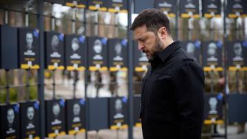 Wołodymyr Zełenski stoi przed ścianą z portretami poległych żołnierzy.