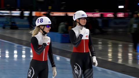 Dwie łyżwiarki w białych kaskach i czerwono-czarnych strojach stoją na lodowisku.