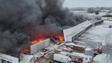 Płonąca hala magazynowa z kłębami czarnego dymu, widoczne wozy strażackie i śnieg na ziemi.