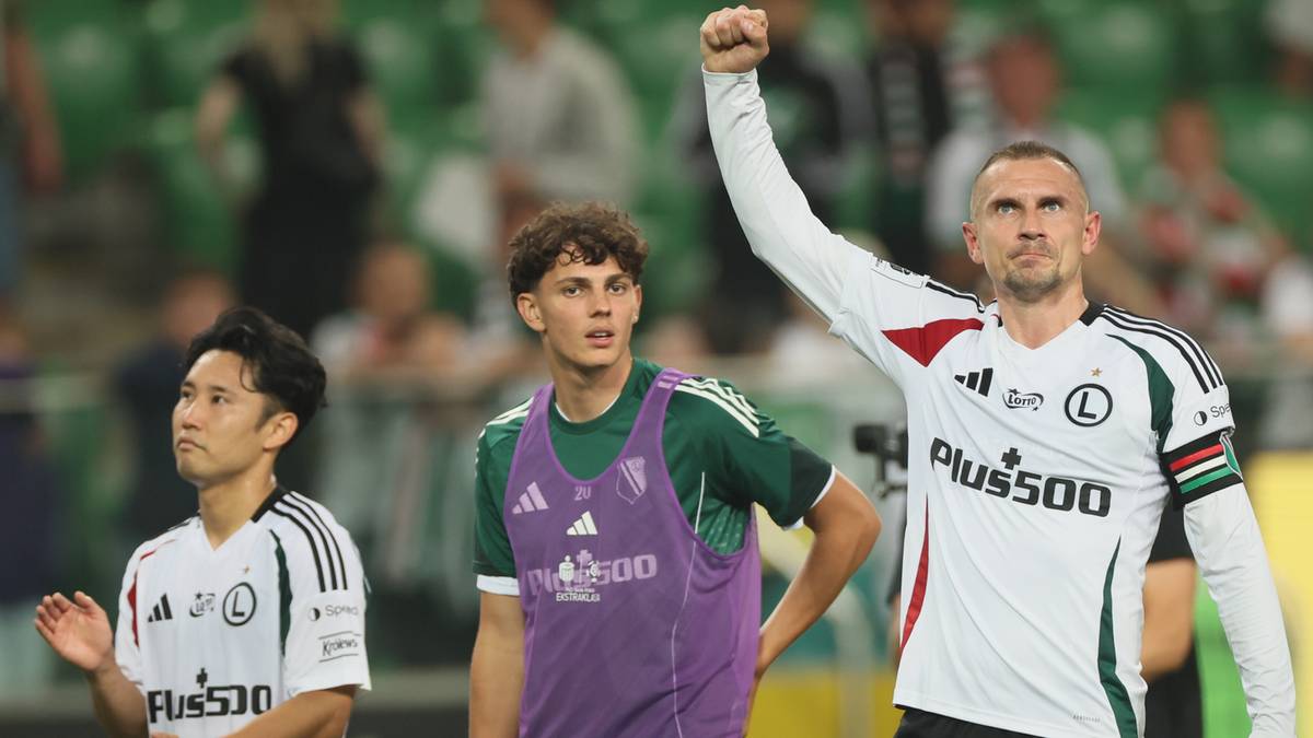 Trzech piłkarzy na boisku, dwóch w białych strojach z czarnymi detalami, jeden w zielonej kamizelce treningowej. Jeden z piłkarzy w białym stroju unosi prawą rękę w geście triumfu.