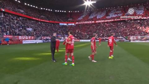 Piłkarze w czerwonych strojach stoją na zielonej murawie stadionu, w tle widać trybuny wypełnione kibicami w czerwonych barwach.