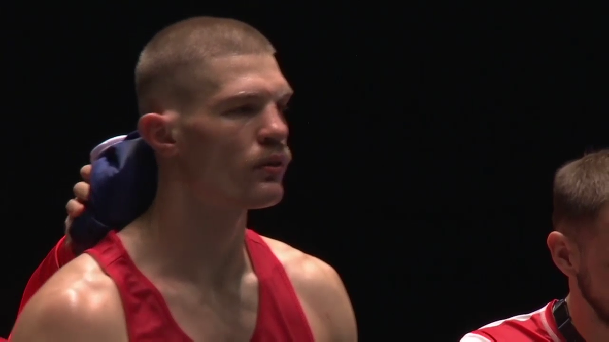 Zbliżenie na twarz młodego boksera w czerwonym stroju, z krótką fryzurą i zamyślonym wyrazem twarzy. W tle widać fragmenty innych osób w strojach sportowych oraz ciemne tło.