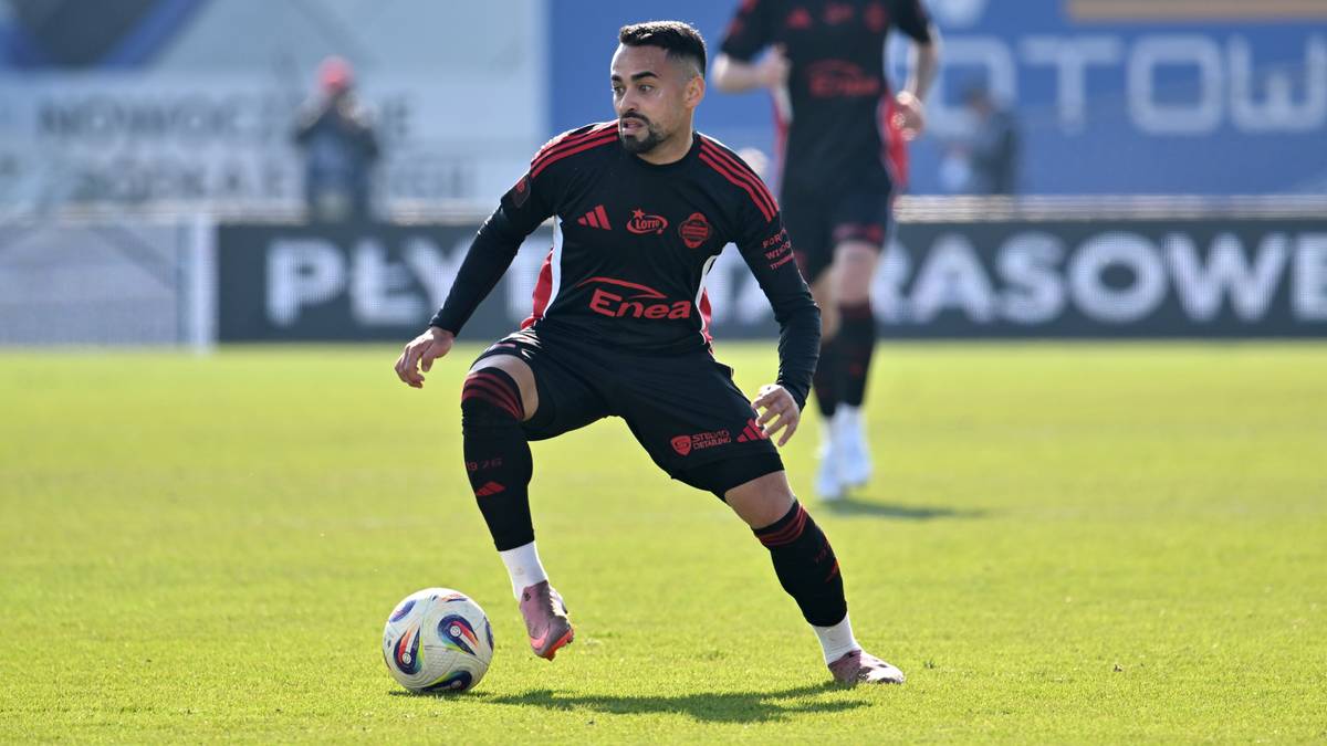 Piłkarz w czarnej koszulce z czerwonymi detalami i czerwonymi spodenkami, prowadzi piłkę po zielonej murawie.