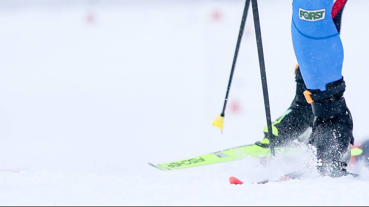 Biathlonista w niebieskim stroju z numerem 7 na piersi biegnie po trasie, trzymając kijki.