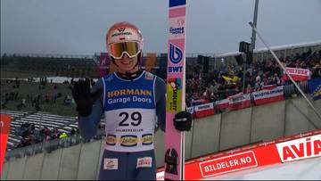 Skoczek narciarski w kasku i goglach, ubrany w niebiesko-szary kombinezon z numerem 29, pozuje do zdjęcia ze swoją różową nartą. Uśmiecha się i macha ręką do widowni zgromadzonej na trybunach.