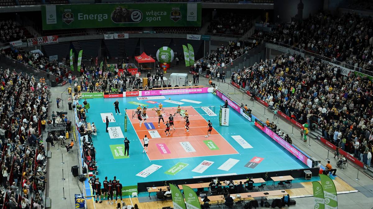 Arena sportowa z meczem siatkówki i liczną publicznością na trybunach.