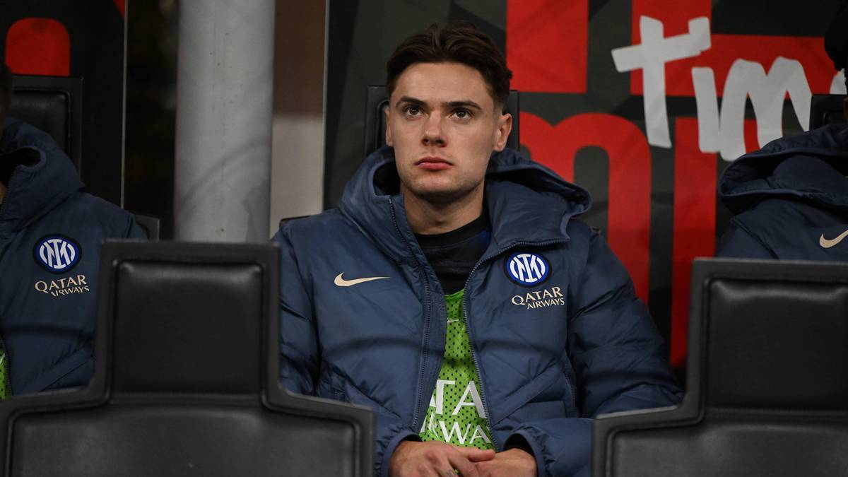 Młody mężczyzna siedzi na ławce rezerwowych w ciemnoniebieskiej kurtce z logo Interu Mediolan i napisem "Qatar Airways". Ma na sobie zieloną koszulkę. Tło stanowi czarna ściana z dużym, białym napisem.