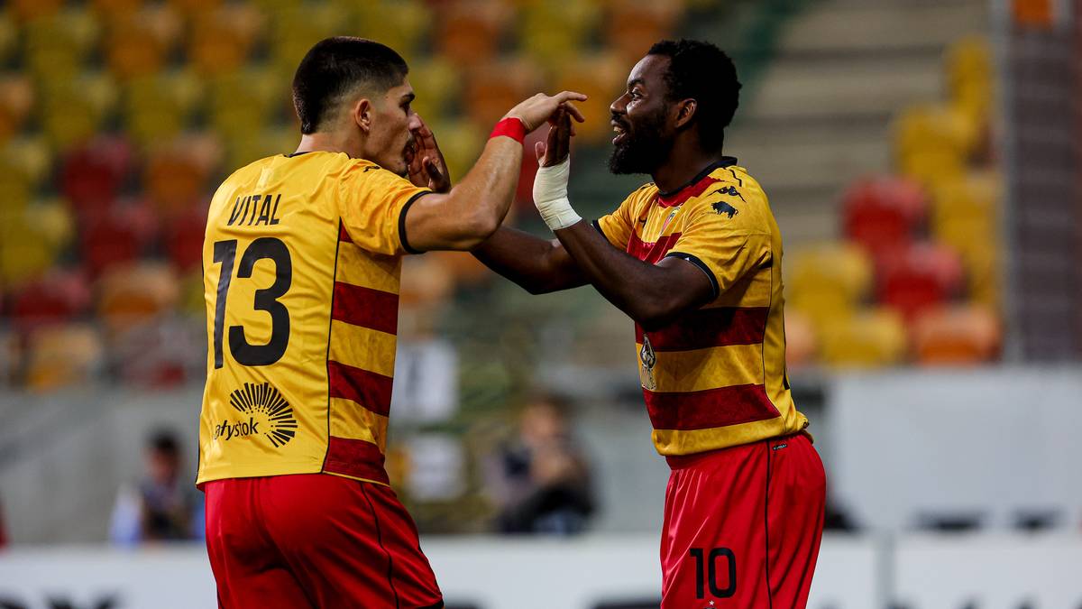 Dwóch piłkarzy Jagiellonii Białystok w żółto-czerwonych strojach podaje sobie ręce na środku boiska, w tle trybuny wypełnione kibicami.