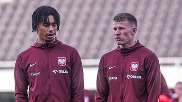 Dw&oacute;ch piłkarzy w bordowych dresach z herbem Polski na piersi stoi na boisku.