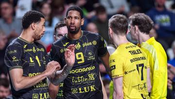 Siatkarze w czarnych strojach z ż&oacute;łtymi akcentami rozmawiają na boisku.