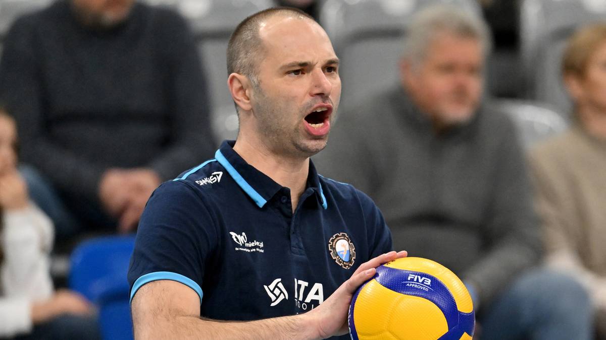 Trener siatk&oacute;wki w ciemnej koszulce sportowej z piłką w dłoni, krzyczący do zawodnik&oacute;w podczas meczu.