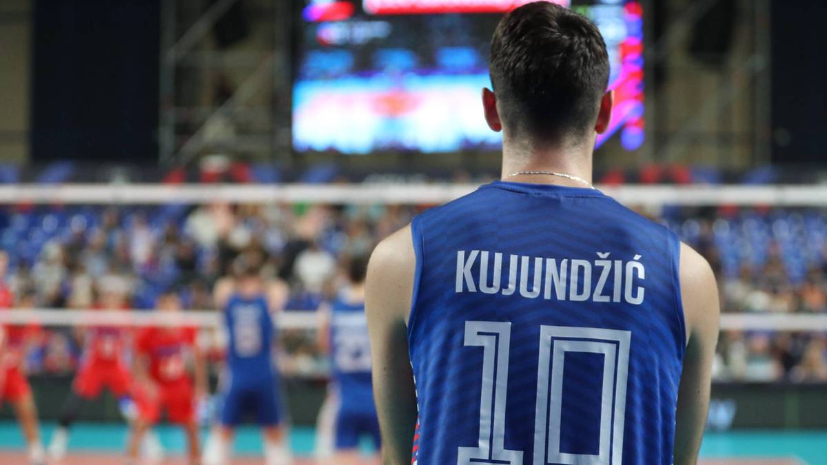 Gracz siatkówki z numerem 10 na plecach, KUJUNDŽIĆ, patrzy na boisko podczas meczu. W tle widoczni inni zawodnicy i trybuny.