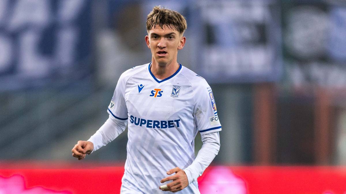 Piłkarz w białym stroju z numerem 27 na plecach prowadzi piłkę po boisku.