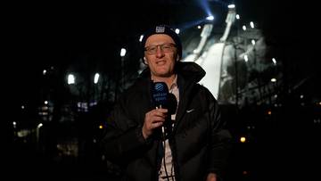 Mężczyzna w czapce z daszkiem i okularach, ubrany w kurtkę, trzyma mikrofon z logo Polsat Sport. W tle widoczna jest nocna scena z reflektorami oświetlającymi konstrukcję przypominającą skocznię narciarską.