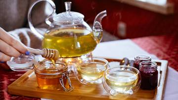 Dłoń nalewa miód z słoiczka do filiżanki na tacy z dzbankiem i innymi naczyniami.