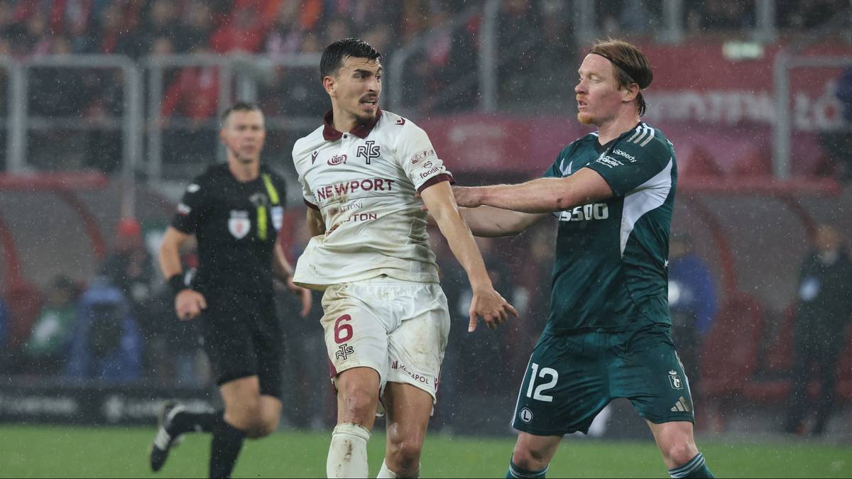 Dwóch piłkarzy walczy o piłkę w powietrzu podczas meczu, sędzia obserwuje akcję w tle, trybuny wypełnione kibicami.