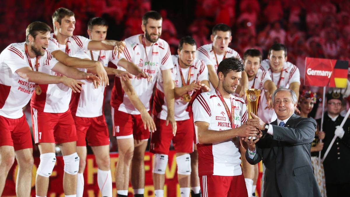 Drużyna siatkarzy w białych koszulkach z czerwonymi elementami, trzymająca złote trofeum, wznosi je do góry podczas ceremonii wręczenia nagród.