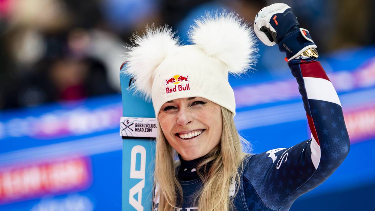 Lindsey Vonn w białej czapce z dwoma białymi pomponami i logo Red Bull, z narzuconą na ramię niebieską nartą z białymi napisami. Kobieta uśmiecha się szeroko, ma zaciśniętą prawą pięść uniesioną do góry.