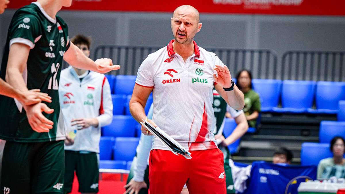 Trener reprezentacji Polski w siatkówce, ubrany w białą koszulkę z czerwonymi elementami i czerwone spodenki, z tabliczką w rękach, przemawia do zawodników.