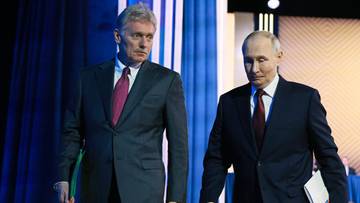 Dmitrij Pieskow i Władimir Putin idący obok siebie w eleganckich garniturach na tle niebieskich zasłon.