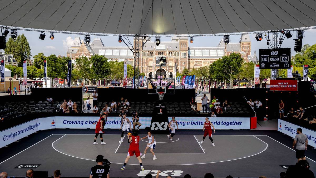 Gracze w czerwonych i białych strojach rozgrywają mecz koszyk&oacute;wki 3x3 na odkrytym boisku, otoczonym trybunami i widoczną architekturą w tle.
