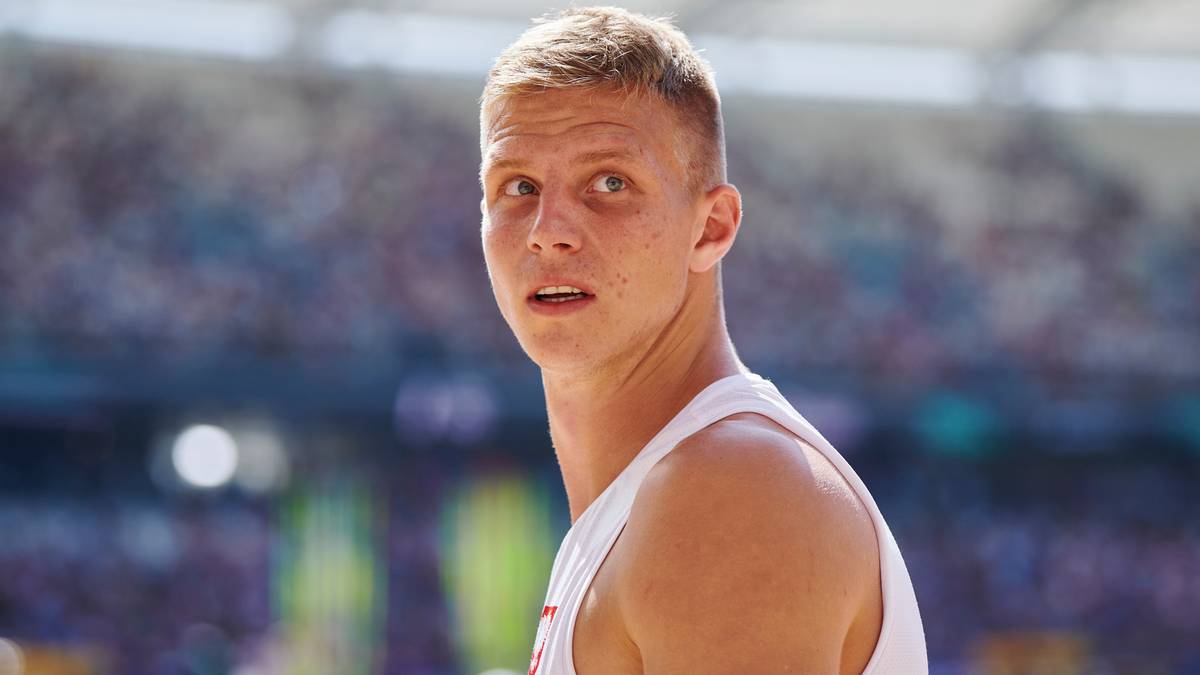 Młody mężczyzna o blond włosach i kr&oacute;tkiej fryzurze, ubrany w białą koszulkę sportową, patrzy w bok z lekko otwartymi ustami. W tle rozmyte trybuny stadionu.