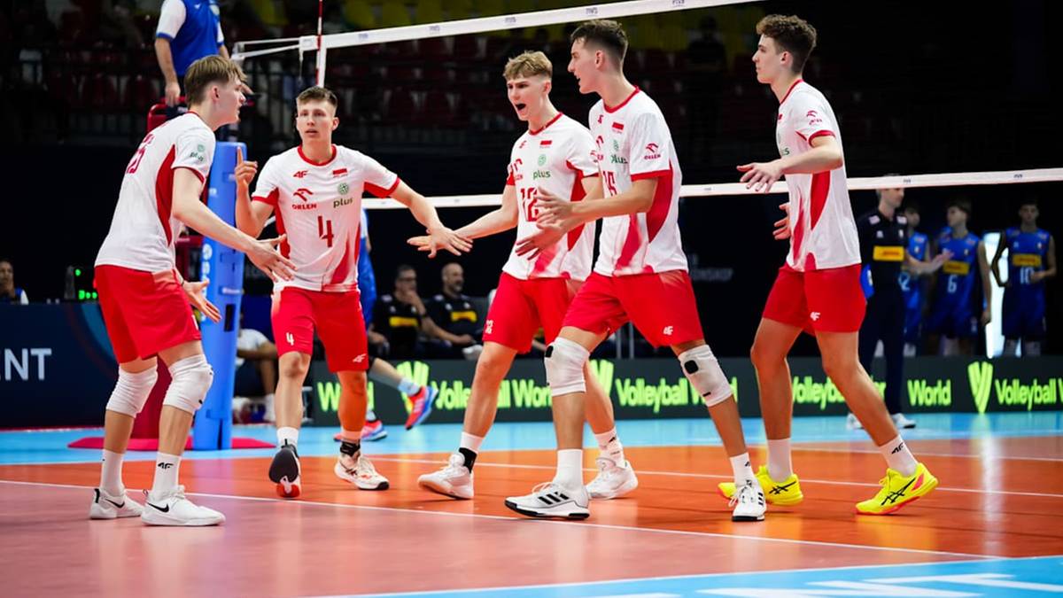 Pięciu młodych siatkarzy w białych koszulkach z czerwonymi elementami i czerwonymi spodenkami stoi na boisku do siatkówki, patrząc na siebie i gestykulując.