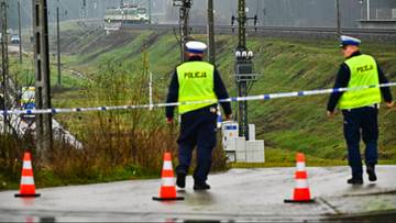 Dwóch policjantów w kamizelkach odblaskowych zabezpiecza teren wokół uszkodzonych torów kolejowych. W tle widać pociąg i infrastrukturę kolejową.