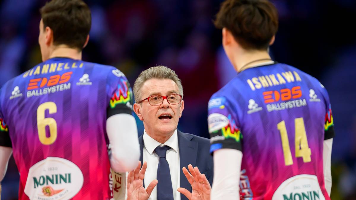 Trener w garniturze przemawia do trzech siatkarzy w kolorowych strojach sportowych.