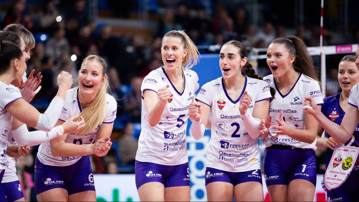 Grupa uśmiechniętych siatkarek w białych koszulkach z niebieskimi elementami, patrzących w stronę widza.