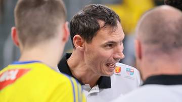 Mężczyzna w średnim wieku, ubrany w sportową koszulkę z kołnierzykiem, z otwartymi ustami i wyrazistą miną, zwraca się do dwóch innych osób w tle. Jeden z nich jest odwrócony tyłem, drugi widziany z profilu. Zdjęcie sugeruje rozmowę lub instruktaż w trakcie meczu sportowego.
