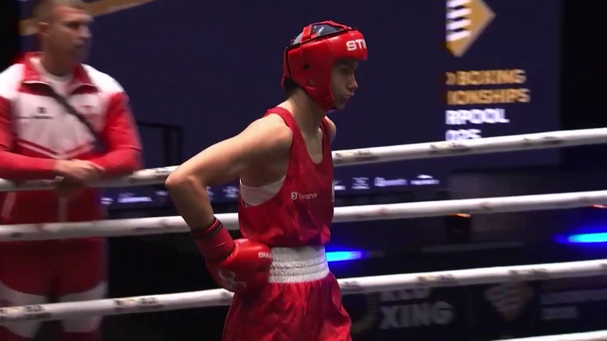 Młoda kobieta w czerwonym stroju bokserskim i kasku stoi na ringu podczas zawodów.