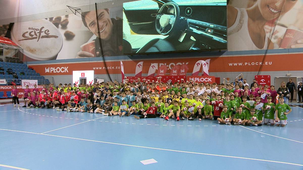 Grupa dzieci w strojach sportowych siedzi na środku boiska hali sportowej.