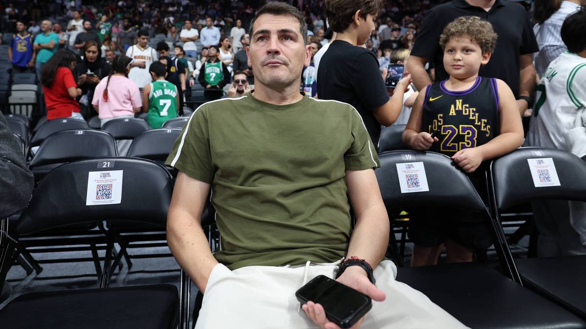 Iker Casillas siedzi na trybunach podczas meczu, trzymając w ręku telefon komórkowy. Obok niego siedzi chłopiec.