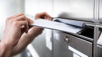 Dłoń wkłada list do metalowej skrzynki pocztowej.