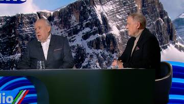 Dw&oacute;ch starszych mężczyzn w garniturach siedzi za stołem, na tle malowniczych ośnieżonych g&oacute;r, prawdopodobnie podczas dyskusji o sporcie.