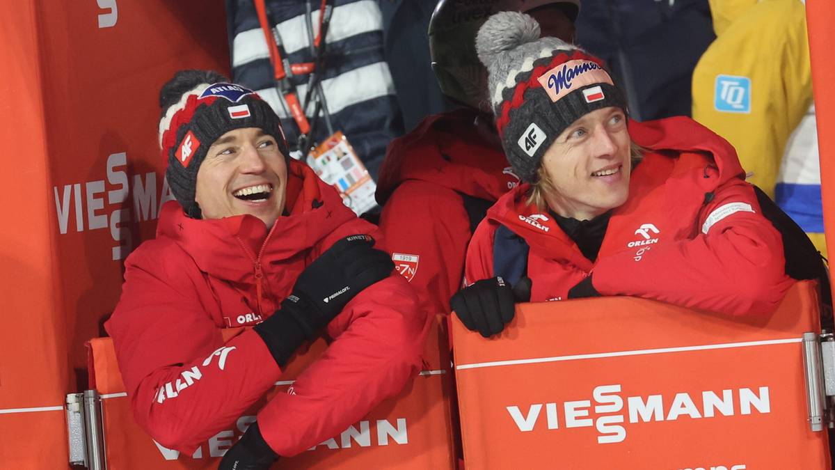 Dwóch skoczków narciarskich w czerwonych kurtkach i czapkach z polskimi symbolami, uśmiechają się.