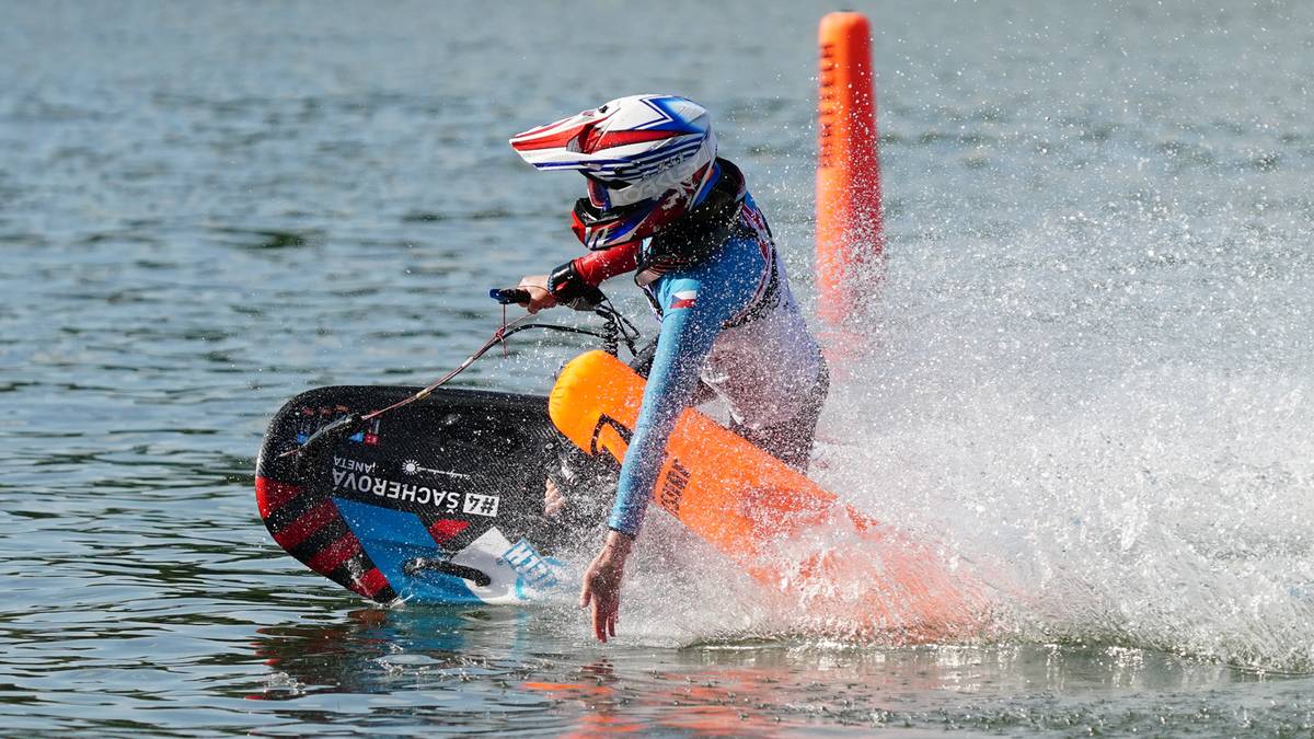 Zawodnik motosurfingu w kasku i kombinezonie pokonuje zakręt w pobliżu pomarańczowych boj, tworząc dużą falę.